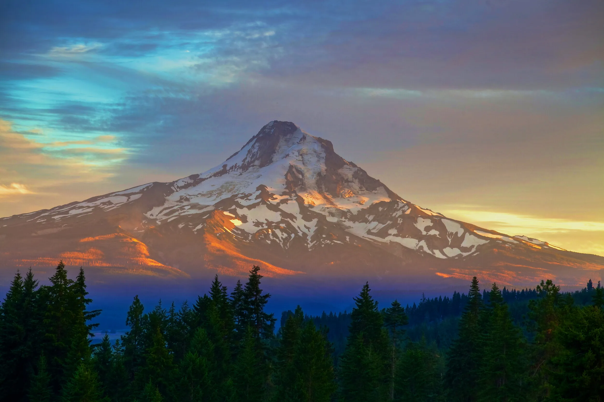 Mount Hood at sunset, its snow-covered peak glowing above a dark ridge of evergreens.